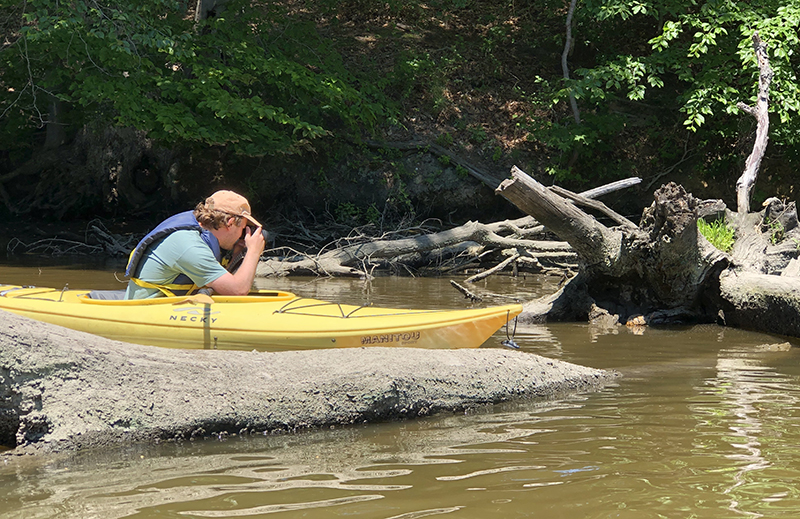 Photographer in kayak photographing something among the branches of a fallen tree by the river.