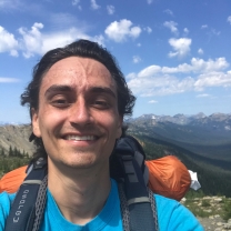 A man wearing a backpack and T-shirt smiles and takes a selfie in a scenic mountain landscape