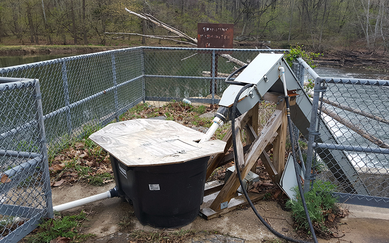 Image of a large tub covered by plywood. The tub is sitting on a concrete slab with a chain link fence on the perimeter. The Patapsco River is in the distance