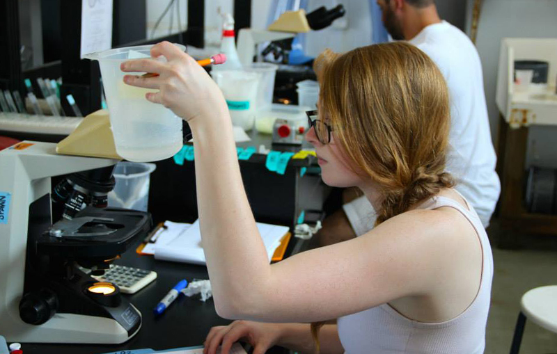 Image of Brittany Wolfe sitting in a lab, holding up a plastic container. A microscope is in front of her.