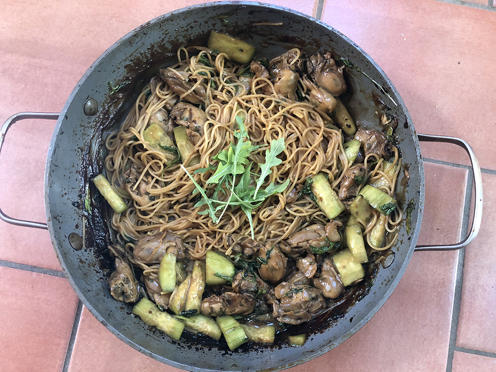 An above image of a wok full of cooked noodles, mussels, and vegetables sitting on a brick floor.