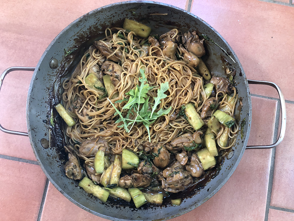 An above image of a wok full of cooked noodles, mussels, and vegetables sitting on a brick floor.