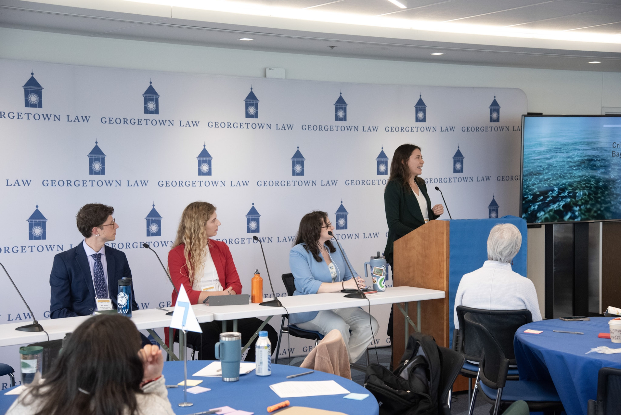 A law student stands at a podium next to a panel of other students and presents her research during a symposium
