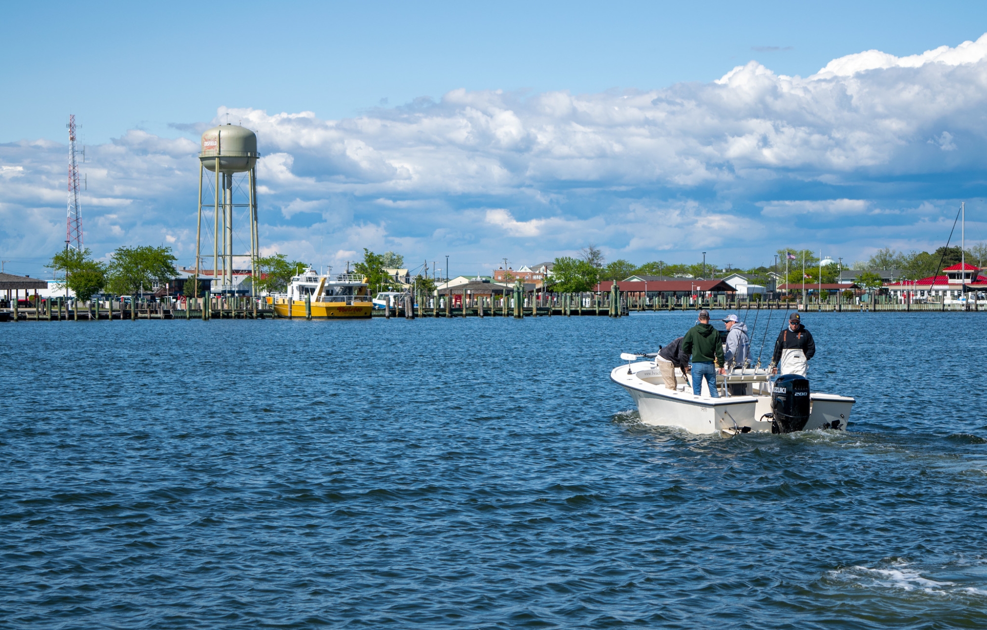 People stand on a boat in the water near a marina with piers and other docked boats 