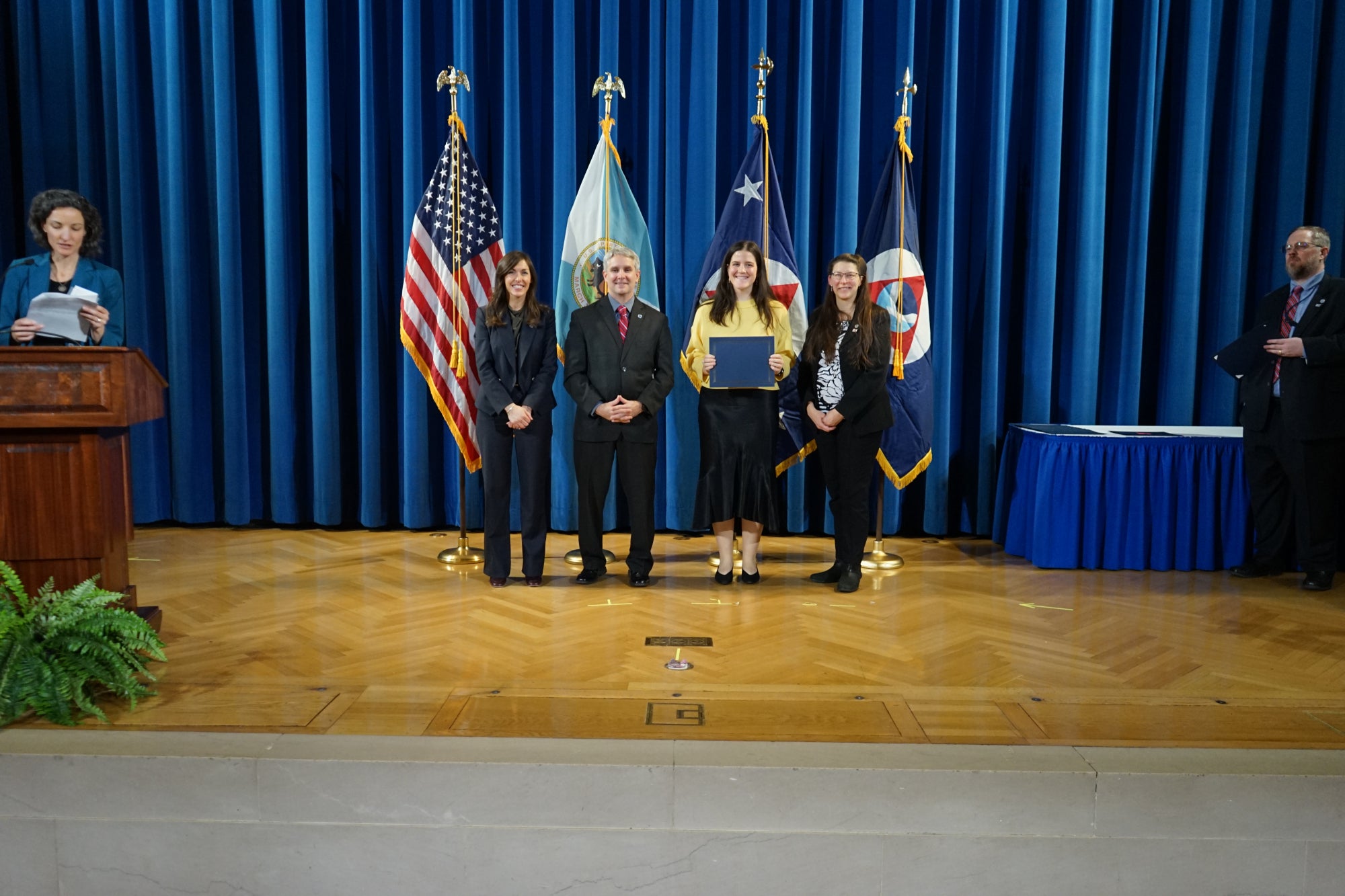 A young woman poses with three people in suits standing in front of flags during a graduation ceremony