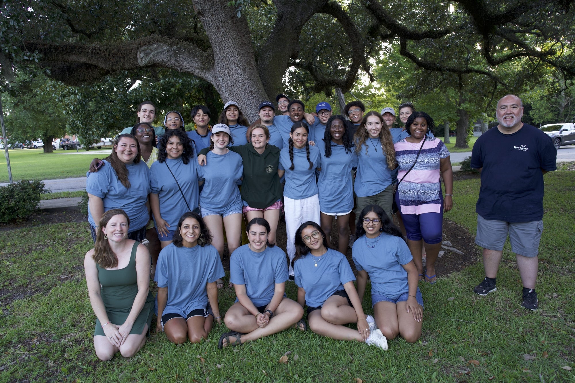 A group of students pose for a photo on a grassy lawn in front of a tree