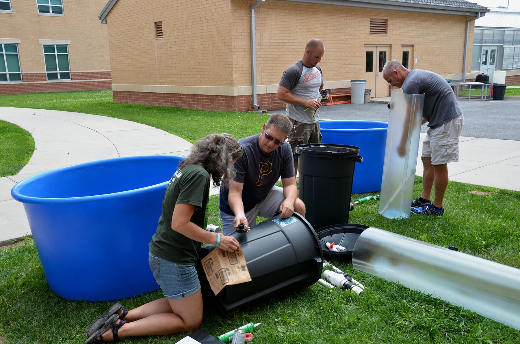 A group of teachers on a lawn outside a school build an aquaculture setup with plastic trash bins, large circular fish tanks, and clear tubes