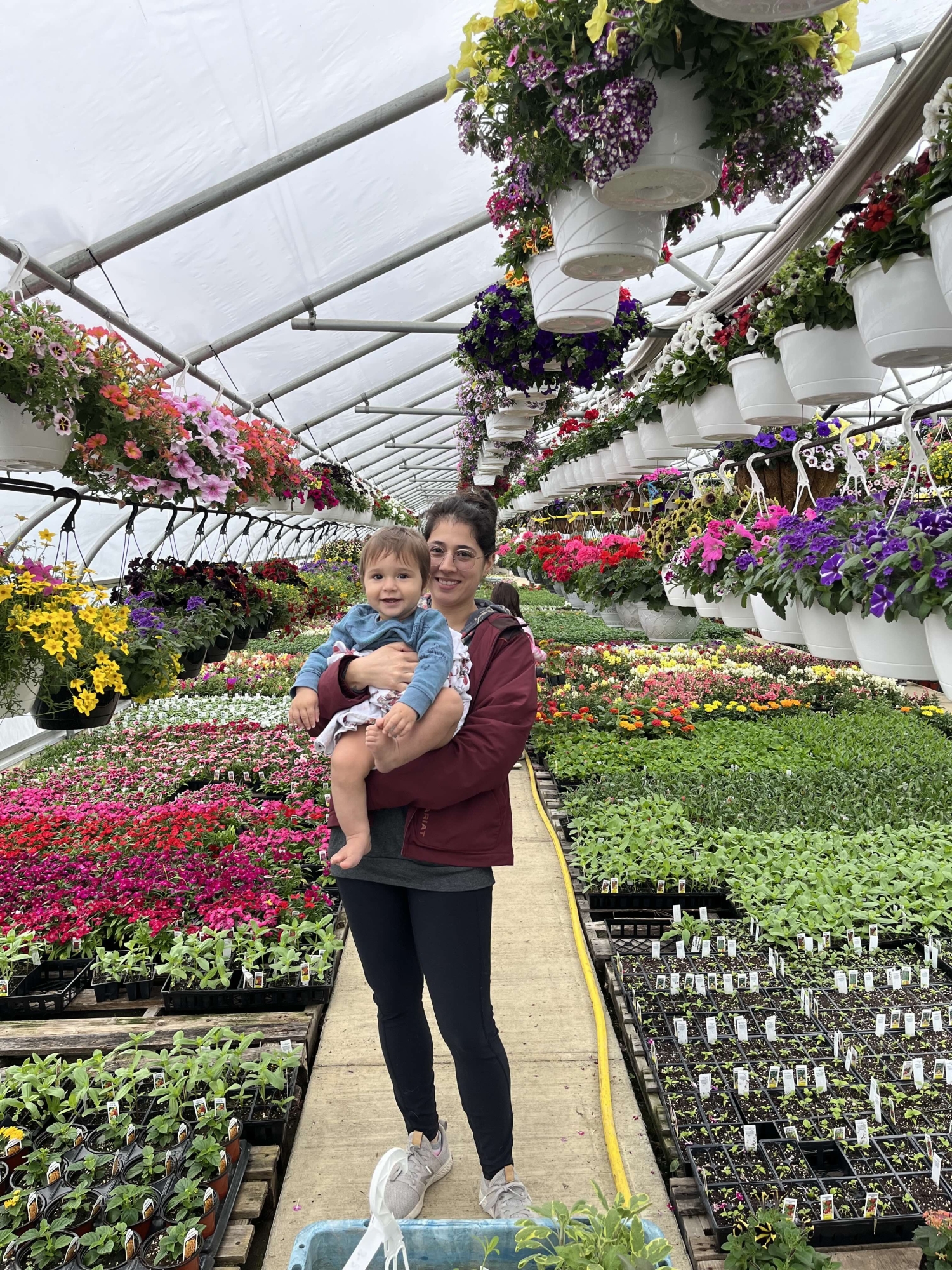 A smiling woman holds her toddler in a greenhouse surrounded by pallets of vegetables and flowers and hanging flower pots