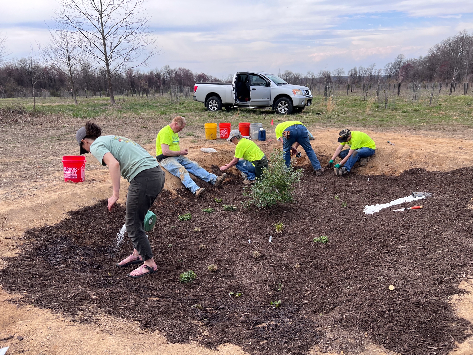 Several people sit around the edges of a garden, planting small plants and spreading mulch around its corners.