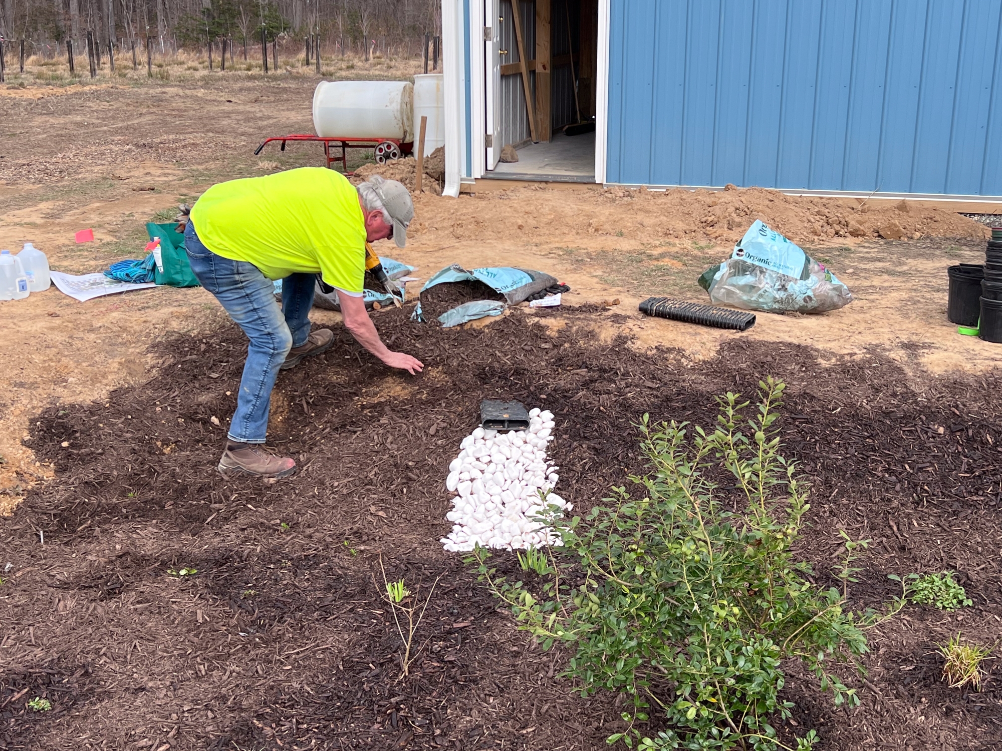 A man pats mulch on the ground near a pile of stones beneath a drainage pipe.