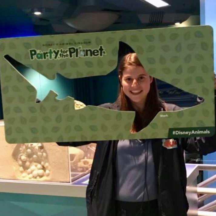 Woman holds up a large green cardboard frame shaped like a fish that reads #DisneyAnimals
