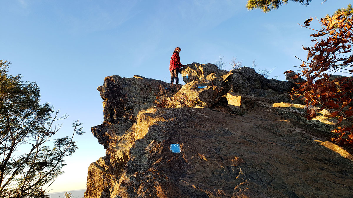 Ashton standing at the top of a steep trail next to a cliff, the height of a tree. Clear sky is in the background.