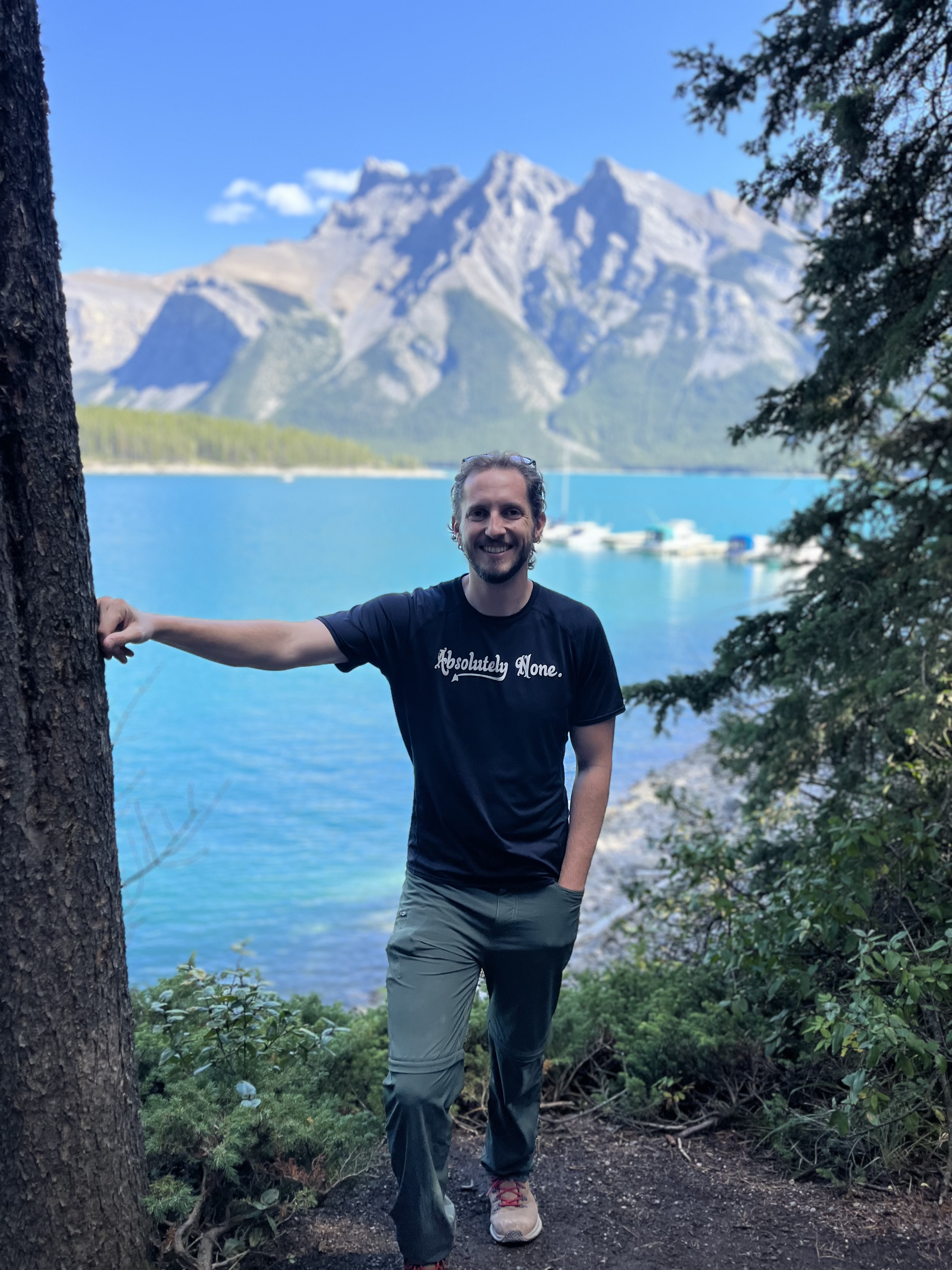 A man wearing a T-shirt and pants poses in front of a large lake with mountains in the background