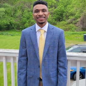 Kai is wearing a navy suit and yellow tie standing on a porch in front of a green yard with trees.
