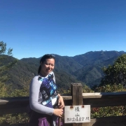 Woman with black hair in ponytail standing in front of mountain view, wearing grey and blue long-sleeve shirt.