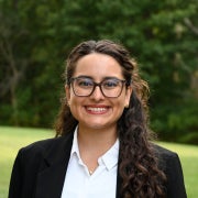 Headshot of Renata smiling wearing a black blazer with trees in the background