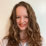 Headshot of a woman with long, wavy brown hair smiling and wearing a grey and white vertically striped, collared shirt.