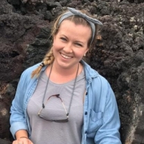 A headshot of Megan Munkacsy, facing the camera smiling, wearing a grey t-shirt with a blue longsleeve shirt open over it