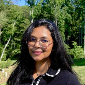 Woman (Syeda Sadia Ali) in a collared blouse and glasses standing outdoors near trees and grass