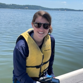 Shayna Keller smiling wearing sunglasses and a yellow life vest, sitting on the edge of a structure with a body of water behind them.