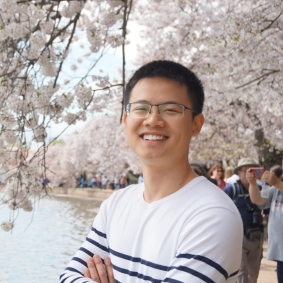 Man wearing white and black striped shirt, smiling and standing in front of cherry blossoms.