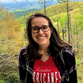 Brunette woman wearing glasses, red shirt and smiling in front of a forest background.