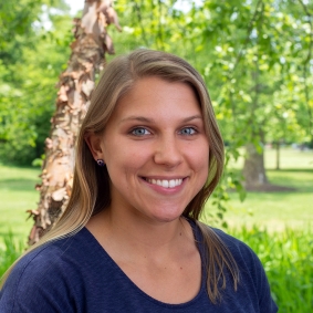 Blonde woman smiling and wearing navy blue shirt in front of green trees.