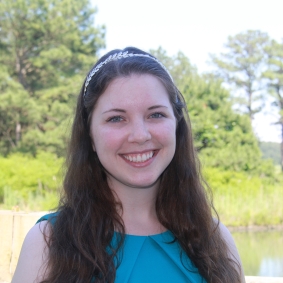 White woman with long brown hair smiling wearing a teal shirt in front of greenery.