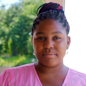 Black woman wearing light pink shirt in front of greenery.