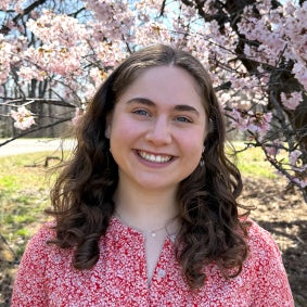 Headshot of a woman with brown curly hair in a red shirt standing in front of a cherry blossom tree.