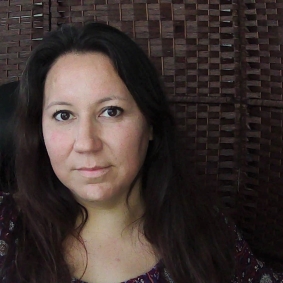 Headshot of a woman with long, dark brown hair and a purple shirt in front of a brown background.