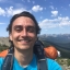 A man wearing a backpack and T-shirt smiles and takes a selfie in a scenic mountain landscape