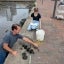 A young woman kneels beside the water in Baltimore's inner harbor with a notepad in hand. A man pulls biofilm discs from the water and lays them on the concrete and brick path