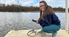 A woman crouches on the end of a boardwalk near a body of water with a handheld monitoring tool attached to a wire