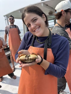 A young woman smiles while wearing waders and holding a large crab.