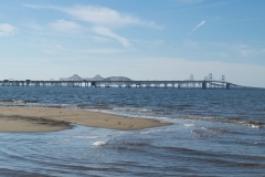 A sandy shoreline of the Chesapeake Bay waters with the Bay Bridge visible in the distance