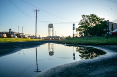 Water pools on a gravel road between a parking lot and building on a sunny day. An old water tower and power lines are in the distance, reflected in the pool of standing water.