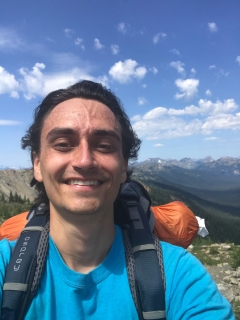 A man wearing a backpack and T-shirt smiles and takes a selfie in a scenic mountain landscape