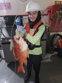 Aiman Raza wears a vest and hard hat and holds a red snapper aboard a research vessel