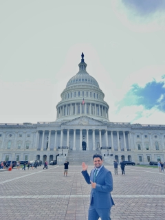 A young man in a suit stands in front of the US Capitol building