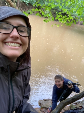 Two people in rain jackets pose for a picture next to a river while taking a water sample in the rain.