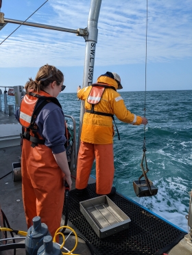 Two researchers wearing waders and life jackets stand on the edge of a boat on the water and lower a tool called a benthic Ponar grab into the water 