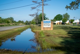 Water stands in a drainage ditch between a school and a road. The school's sign can be seen.