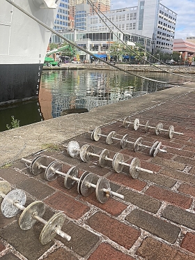 Five poles with circular disks attached to them are laid out on a brick pathway next to water in Baltimore's Inner Harbor