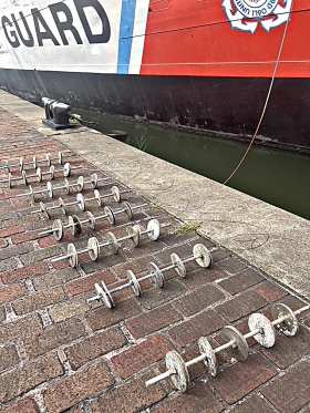 A series of biofilm racks (poles with circular disks attached) laid on a brick pathway next to a ship in Baltimore's Inner Harbor