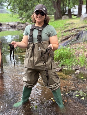 Julia Devine stands in a shallow creek wearing waders and holding long, stringy mats of algae