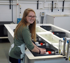 Image of Brittany Wolf standing by an oyster seed tank and looking over her right shoulder toward the photographer