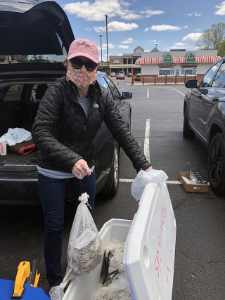 Image of woman in mask by open car trunk in a large mall parking lot. She is holding a bag of oysters that she is putting into a white cooler.