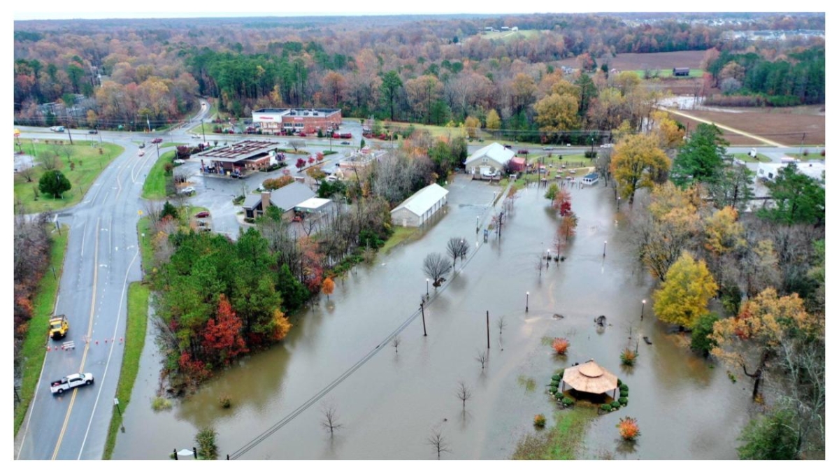 Flooding alongside a road in area with cars, trees, and houses