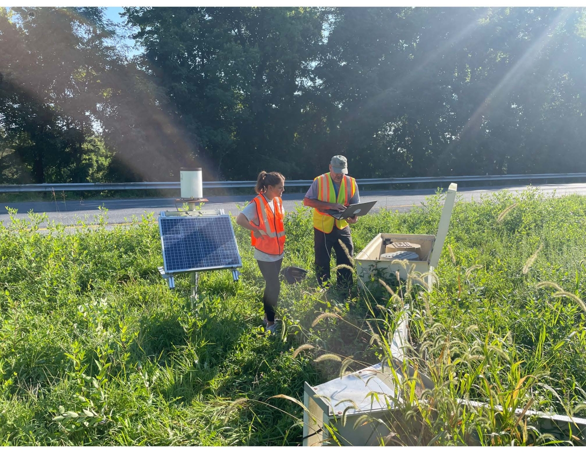 Two researchers stand in a grassy area looking at stormwater mitigation equipment near a roadside. One researcher holds a laptop.